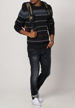 Pull rayé marine avec des lignes rouges et bleu clair, associé à un jean noir et des baskets blanches. Le modèle porte un sac à dos beige.