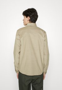 Camisa de manga larga beige con cuello, textura fina y dobladillo en curva. Cuenta con puños de botón. Combinada con pantalones verde oscuro.