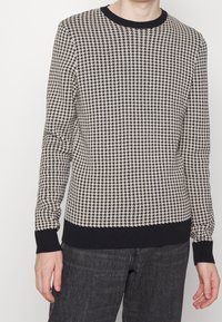 Knit sweater featuring a black and cream houndstooth pattern, with a black neckline and cuffs. Material appears soft with a textured finish.