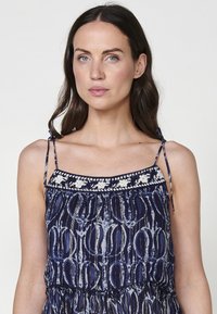 Navyblauwe jurk met dunne bandjes, gestructureerde stof, elastische taille en bloemenborduursel bij de halslijn. Heeft een vloeiende silhouet.