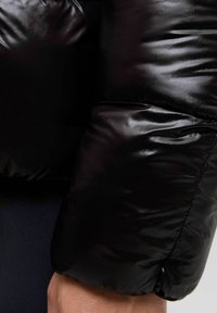 Primo piano di una manica di una giacca a vento nera lucida appoggiata sulla pelle, che mostra la texture del tessuto e i dettagli delle cuciture.