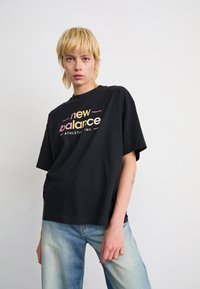 Asmuo su trumpais šviesiais plaukais, dėvintis juodą New Balance marškinėlį ir šviesiai mėlynas džinsus, prieš paprastą baltą foną.