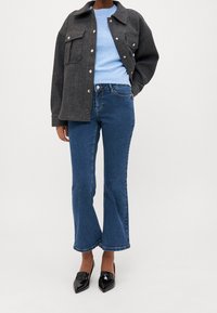 Szara kurtka z mieszanki wełny z zatrzaskami, niebieski sweter dziany, rozszerzane niebieskie dżinsy oraz czarne lakierowane szpilki, prezentujące casualowy, warstwowy look.