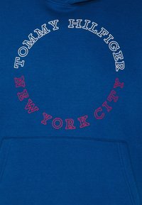 Modré mikina s kapucí s kulatým logem. "TOMMY HILFIGER" je napsáno bílou barvou, "NEW YORK CITY" červenou barvou a obsahuje přední kapsu.