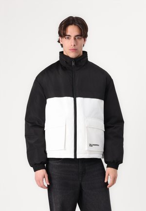 Puffa jakna u crno-bijeloj boji s visokim ovratnikom, zatvaranjem na zip i dva prednja džepa. Ima pletene manžete i brendiranje na rubu.