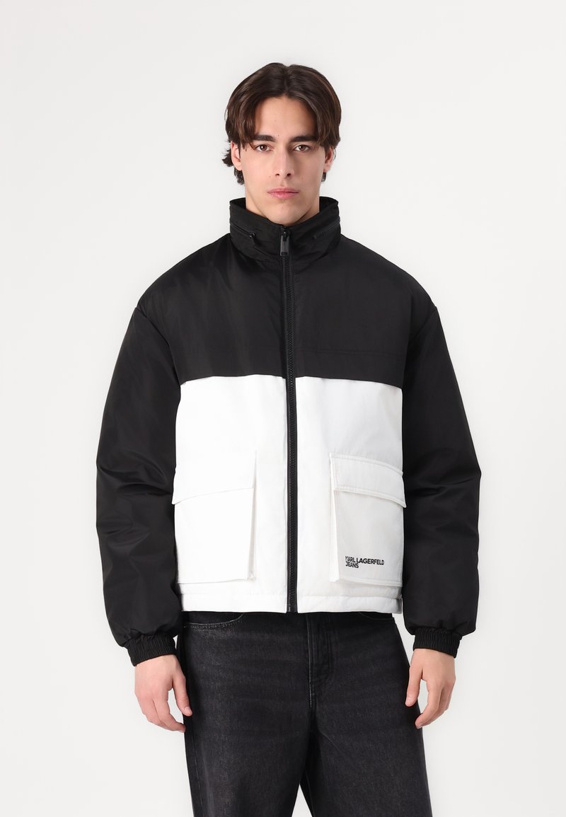 Black and white color-block puffer jacket with a high collar, zip closure, and two front pockets. Features knitted cuffs and branding on the hem.