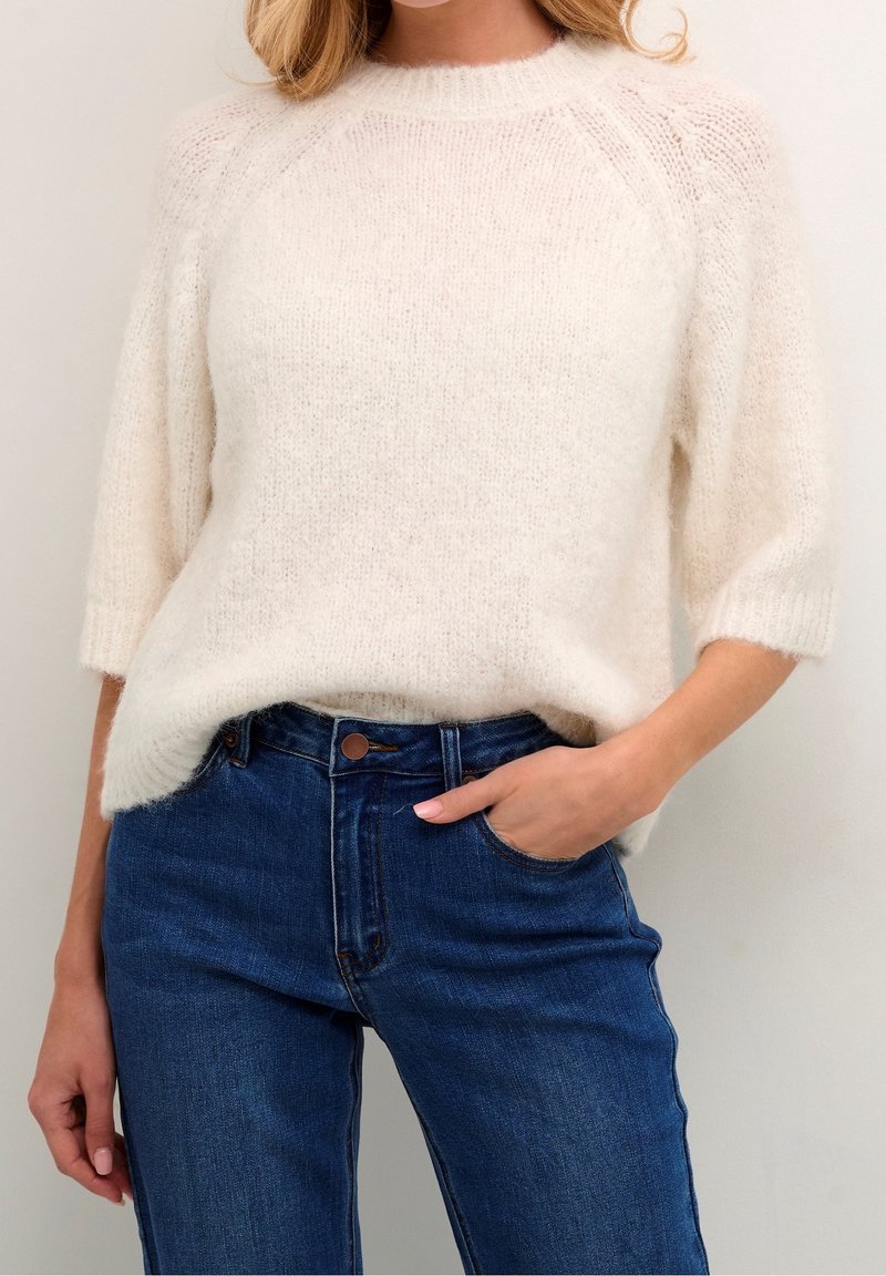 Maglione lavorato a maglia color crema, soffice, con maniche corte e scollo rotondo, abbinato a jeans denim blu scuro. Vestibilità casual con una texture sottile.