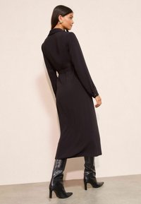 Musta keskmise pikkusega kleit, millel on krae, pikad varrukad ja vöödiga talje. Seljas on põlvikute kõrgused mustad kontsakingad, näidates sujuvat kangast tekstuuri.