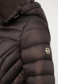 Veste matelassée marron avec col en fausse fourrure, présentant un design matelassé et un accent logo doré sur la manche. Texture lisse et brillante.