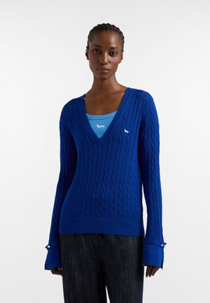 Kvinde iført en blå kabelstrikket V-hals sweater over en lysere blå tanktop med et lille hvidt gravhundlogo, kombineret med mørke bukser.