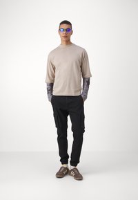 Camiseta beige de manga corta, camisa gris de manga larga debajo, pantalones cargo negros y zapatillas marrones con rayas blancas. Gafas de sol azules.