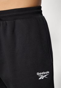Os fatos de treino Reebok pretos apresentam uma cintura canelada, tecido suave e um logotipo branco na parte inferior da perna, destacando um design simples e desportivo.