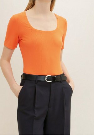 Raitapintainen oranssi t-paita, jossa on kulmikas kaula-aukko, yhdistettynä korkeavyötäröisiin tummansinisiin housuihin ja mustaan vyöhön, jossa on hopeinen solki.