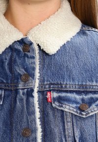 Giacca di denim con colletto in pelliccia sintetica bianca, bottoni in rame e etichetta rossa Levi's. Presenta tasche sul petto anteriori e bordi sfrangiati.