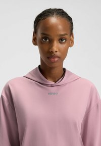 Ljusrosa hoodie med hög krage och HUGO-logotyp i teal. Mjuk textur, avslappnad passform och sänkta axelsömmar.