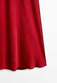 Un long vêtement rouge fluide se drape avec grâce, mettant en valeur un tissu texturé et un ourlet lisse sur un fond blanc.