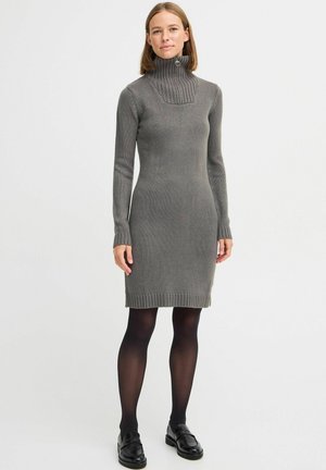 Robe en tricot gris avec un col montant côtelé, des manches longues et une coupe ajustée. Présente une légère évasement à l'ourlet et des poignets côtelés. Chaussures noires.