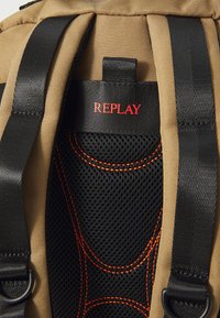 Tan backpack with black mesh padding, black straps, and red stitching. Features a prominent red "REPLAY" logo on the top.