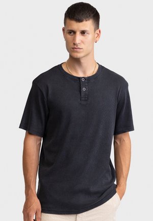 Νέος άνδρας με κοντά σκούρα μαλλιά που φοράει μαύρο κοντομάνικο πουκάμισο Henley με δύο κουμπιά, κοιτάζοντας δεξιά, με τα χέρια στις τσέπες μπεζ παντελονιού.