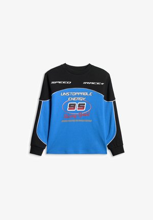 Chłopiec z krótkimi blond włosami, ubrany w czarno-niebieską bluzę z napisem „Unstoppable Energy 95 Racing Speed”, stoi na jednolitym tle.