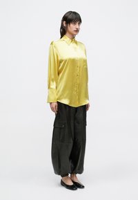 Femeie purtând o cămașă galbenă strălucitoare cu nasturi, pantaloni largi negri în stil cargo cu manșete elastice și pantofi negri comozi, stând în picioare în fața unui fundal simplu.