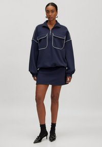 Marineblauwe zip-up sweatshirt met twee grote zakken en witte piping, gecombineerd met een bijpassende strakke rok en zwarte puntige hakken.