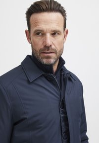 Chaqueta aislante azul marino con un collar estructurado, botones frontales y cierre de cremallera. El material exterior es suave con un acabado ligeramente brillante.