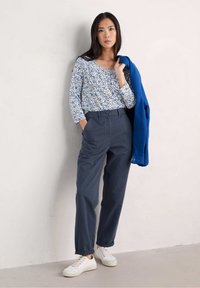 Witte bloemen lange mouwen top met blauwe accenten, donkergrijze broek, witte sneakers en een blauwe jas over de schouder gedrapeerd.