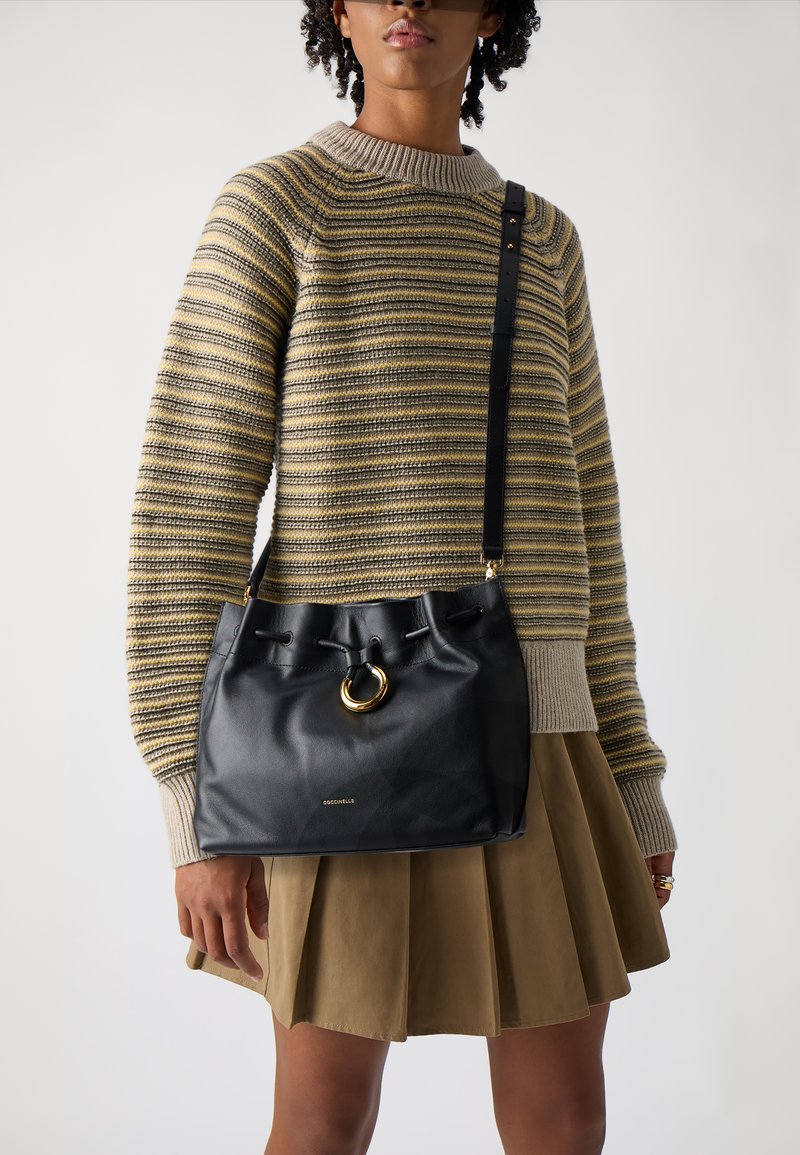 Borsa a secchiello in pelle nera con un elemento ad anello in metallo dorato, chiusura a coulisse e tracolla regolabile, abbinata a un maglione a righe e una gonna.