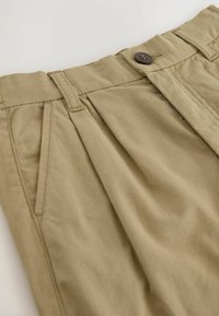 Pantalon couleur kaki avec passants pour ceinture, poches latérales, plis et fermeture par bouton à la taille.