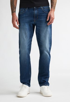 Hombre con vaqueros azules deslavados y zapatillas blancas, de pie contra un fondo liso, con antebrazos tatuados visibles.