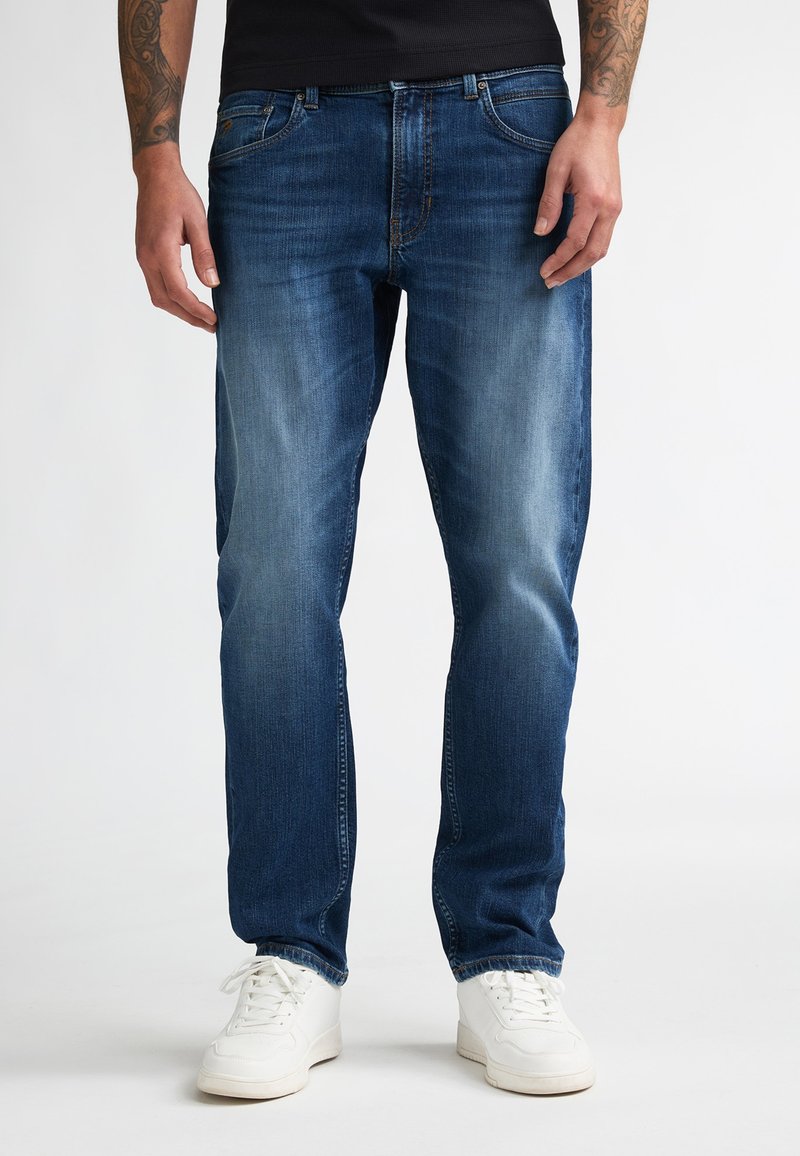 Homme portant un jean bleu délavé et des baskets blanches, debout devant un fond uni, avec les avant-bras tatoués visibles.