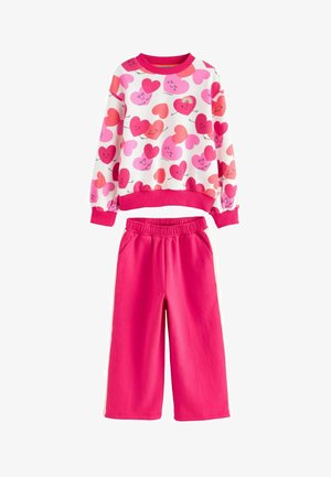 Súprava mikiny a teplákov. Biela mikina s ružovými srdiečkovými vzormi; rebrované manžety. Jasne ružové nohavice s elastickým pásom a bočnými vreckami.