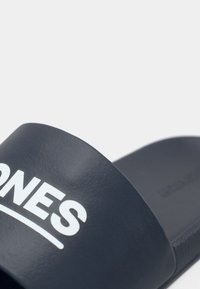 Nahaufnahme eines marineblauen Slides mit weißem Teiltext "ONES" auf dem Riemen, vor einem schlichten weißen Hintergrund.