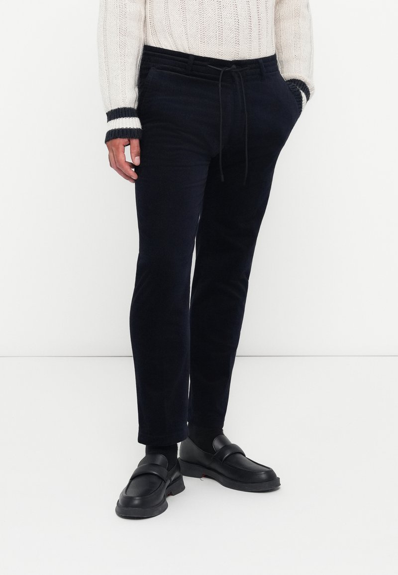 Marineblauwe corduroy broeken met een trekkoord in de taille, voorzien van een slim fit en een soepele textuur. Gecombineerd met zwarte instap schoenen.