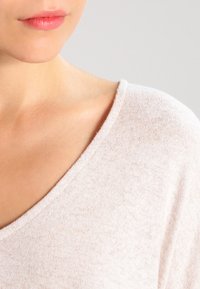 Jasnoróżowy, fakturowany materiał o luźnym kroju i dekolcie w kształcie litery V. Skupia się na obszarze ramion i dekoltu, z wyraźnie gładką fakturą.