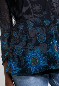 Blusa negra transparente con patrones circulares azules y verdes, combinada con jeans de mezclilla azul claro, mostrando un primer plano de la parte baja del torso y el brazo.