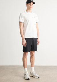 Cămașă polo albă cu margini negre, asortată cu pantaloni scurți negri texturați. Modelul poartă șosete albe și încălțăminte sport gri, completat cu o șapcă neagră.