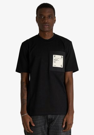 Giovane uomo che indossa una maglietta nera con una grande etichetta quadrata sulla tasca del petto e jeans scuri, in piedi davanti a uno sfondo semplice.