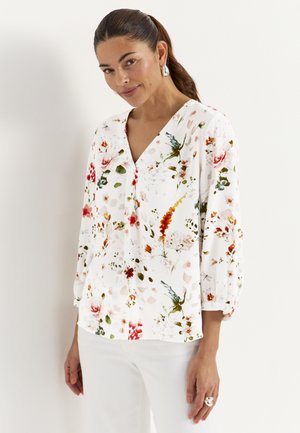 Kobieta o brązowych włosach związanych z tyłu, nosząca srebrne kolczyki, białą kwiecistą bluzkę z czerwonymi, zielonymi i pomarańczowymi kwiatami oraz białe spodnie, stojąca bokiem.