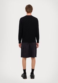 Maglione nero lavorato a maglia con un motivo a diamante testurizzato, abbinato a pantaloni cargo scuri e scarpe nere eleganti, visto da dietro.