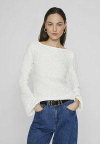 Top bianco a maniche lunghe con texture, scollatura a barca e polsini svasati, abbinato a jeans blu a vita alta e una cintura nera.