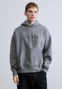 Szürke kapucnis pulóver kenguruzsebbel, fekete "CALVIN NEW YORK" hímzett logóval. Puha, texturált anyagból készült.