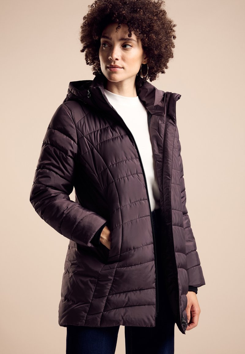 Lila pufferjacka med quiltat mönster, dragkedjestängning och huva. Har sidofickor och en mjuk textur. Bärs över en vit topp.