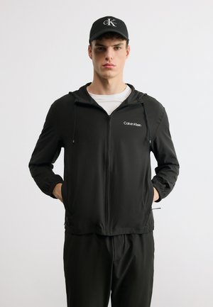 Νέος άνδρας που φοράει μαύρο φούτερ με φερμουάρ Calvin Klein, άσπρο T-shirt, μαύρο παντελόνι που ταιριάζει και μαύρο καπέλο με το λογότυπο CK, στέκεται με τα χέρια στις τσέπες.