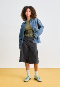 Veste en denim sur une chemise vert olive, associée à un short cargo noir. Des baskets vert clair et des chaussettes grises complètent la tenue.