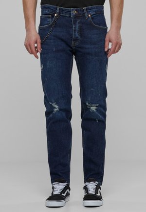 Person iført mørkeblå slidte jeans med en kæde på lommen og sorte sneakers med hvide snørebånd og såler.