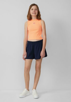 Oranje geribd crop-top met een bloemenembleem, gecombineerd met marineblauwe shorts met elastische taille en witte sneakers. Over het geheel gladde texturen.