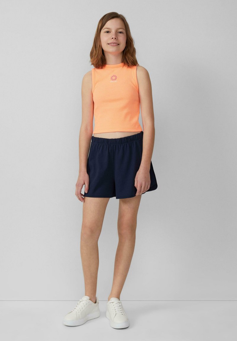 Oranje geribd crop-top met een bloemenembleem, gecombineerd met marineblauwe shorts met elastische taille en witte sneakers. Over het geheel gladde texturen.