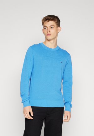 Light blue ribbed knit sweater with a round neckline and long sleeves, featuring a small logo on the chest. Paired with black pants.